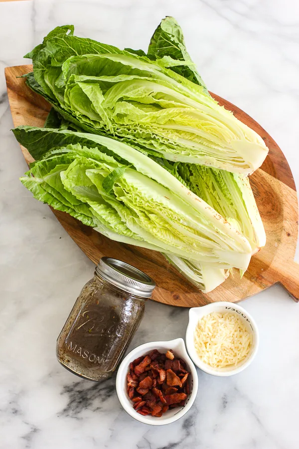 Showing the cut romaine before grilling with the jar of vinaigrette, the cooked bacon bits and the shredded cheese all in separate containers