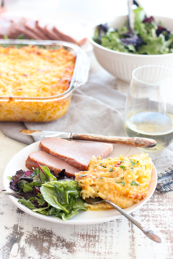 Finished cheesy hash browns plated with sliced ham and salad greens with Serving hash browns serving dish, sliced ham and salad in the background