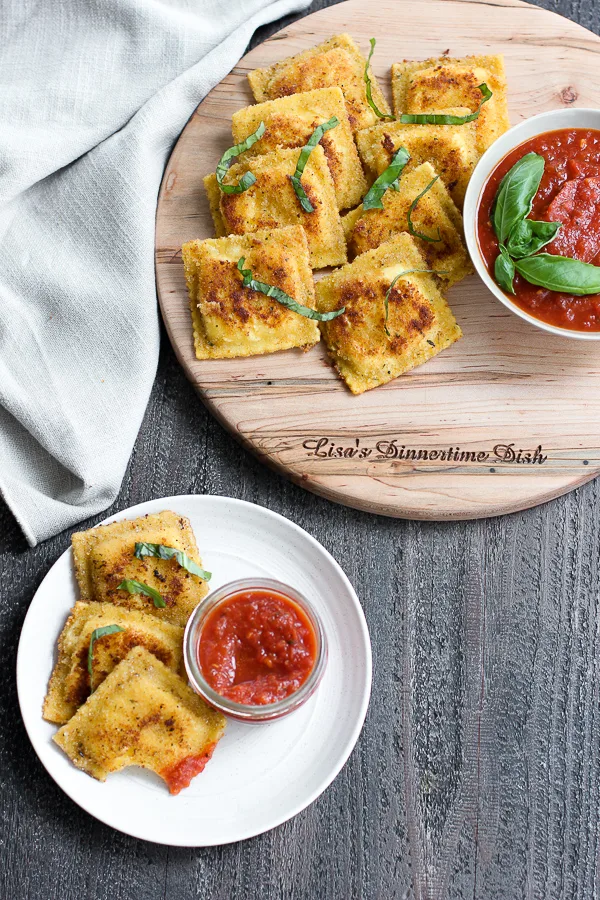 Fried ravioli on a wooden serving board served with marinara sauce and garnished with fresh basil along with a appetizer plate with 3 raviolis and marinara sauce