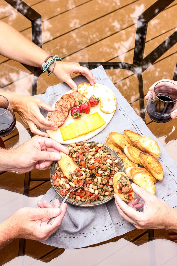 Italian Sausage and White Bean Bruschetta on table showing people topping toasted bread with the sausage and bean mixture and served with wine.