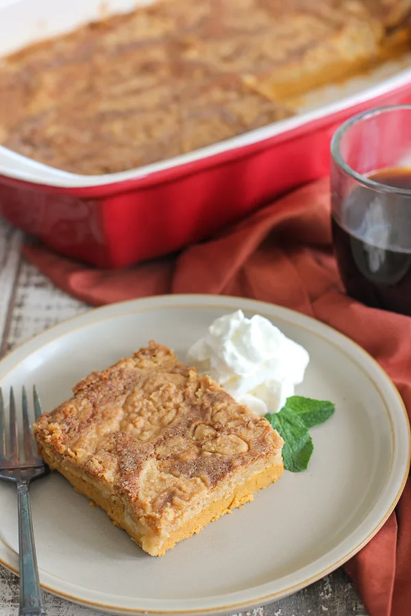 A slice of pumpkin dump cake plated and garnished with whipped cream and fresh mint, served with coffee with the pan of the dump cake in the background