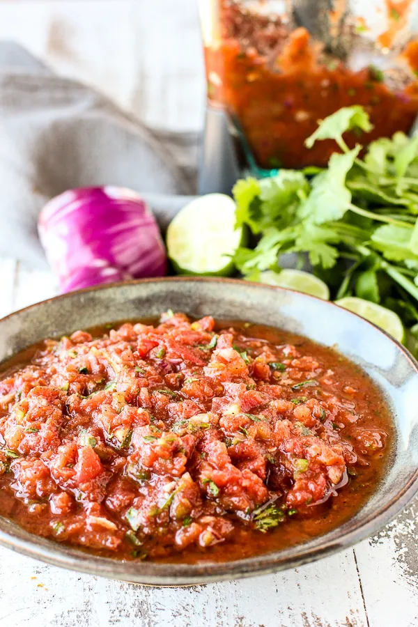Close up of Restaurant Style Salsa in a serving bowl with onion, lime, cilantro and the blender in the background