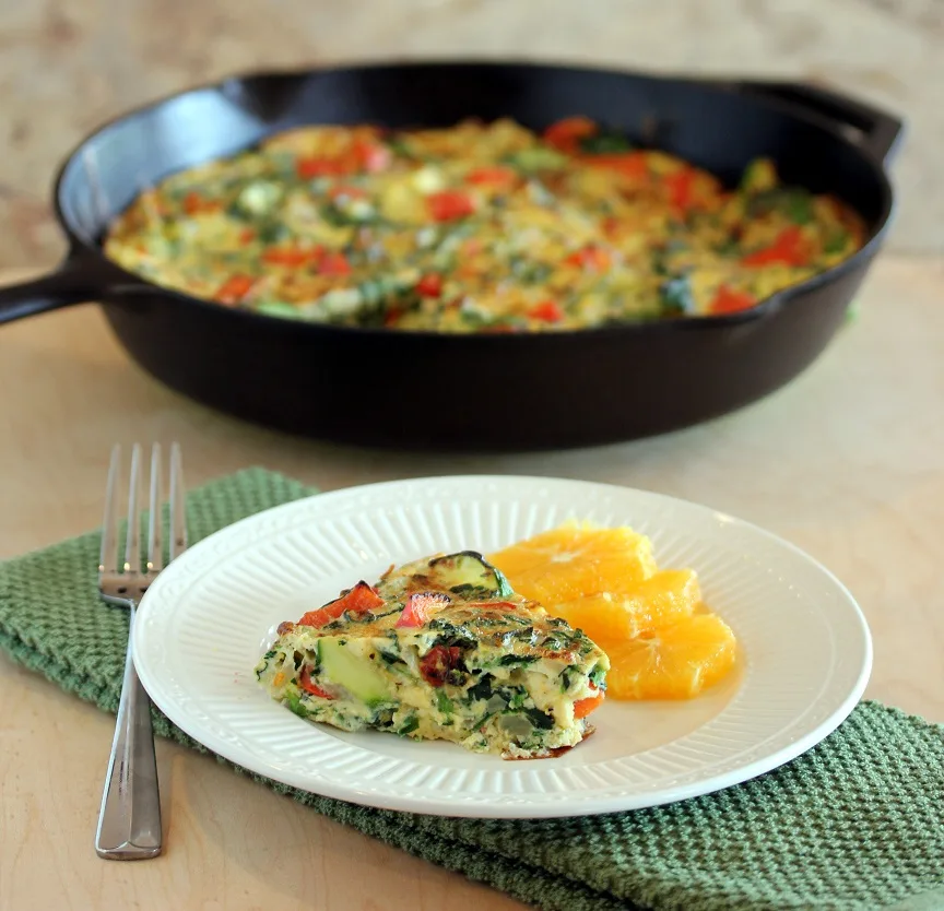 A slice of veggie frittata served with fresh orange slices with the skillet tin the background