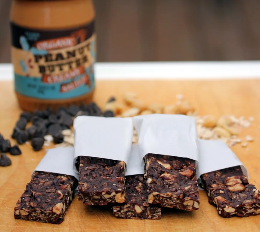 chocolate peanut butter granola bars wrapped in parchment with a jar of peanut butter and loose chocolate chips and oatmeal in the background