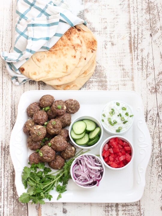 Lamb meatballs with tzatziki sauce shown on a platter with cucumber, red onion and tomato garnishes with pita bread off to the side 
