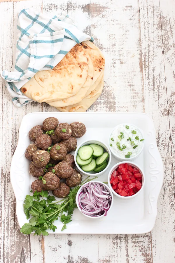 lamb meatballs on a serving platter with onion, cucumber and tzatziki sauce, along with warm naan bread wrapped in a napkin