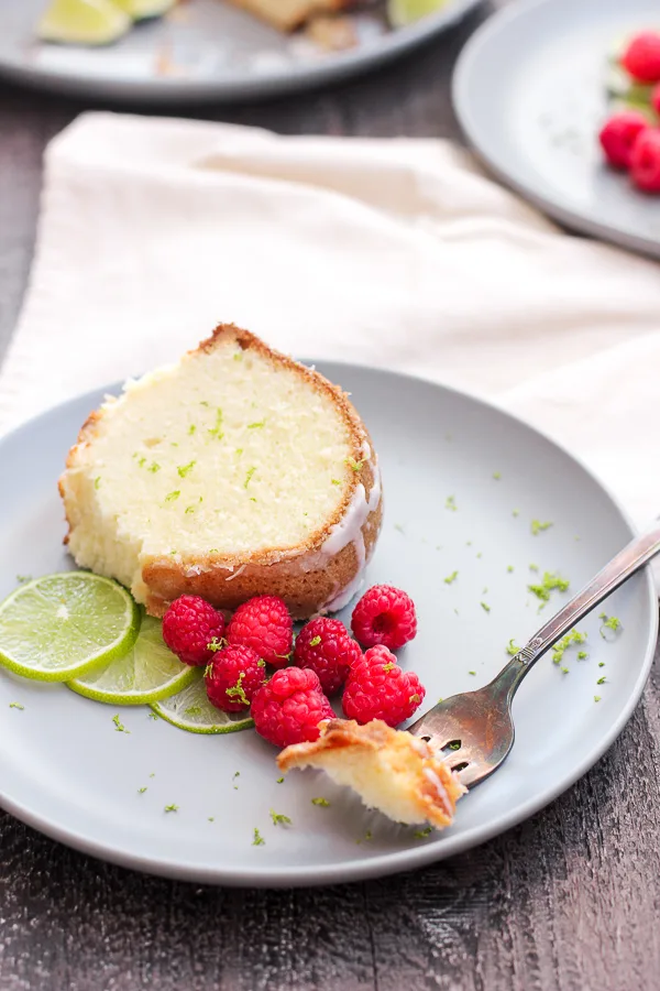 Plated slice of lime pound cake with a bite on a fork and garnished with raspberries and lime slices