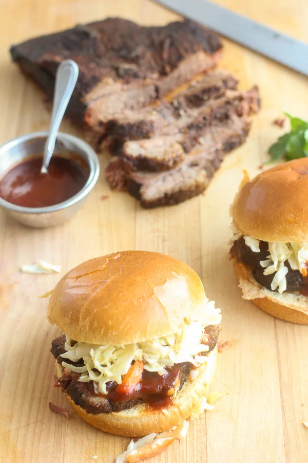 Overhead photo of the sliced brisket served on brioche buns and topped with barbecue sauce and coleslaw withe the sliced brisket in the background along with a dish of barbecue sauce