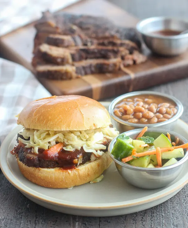 Slow cooker brisket sandwich served with cucumber salad and baked beans, with sliced brisket in the background on a wooden board