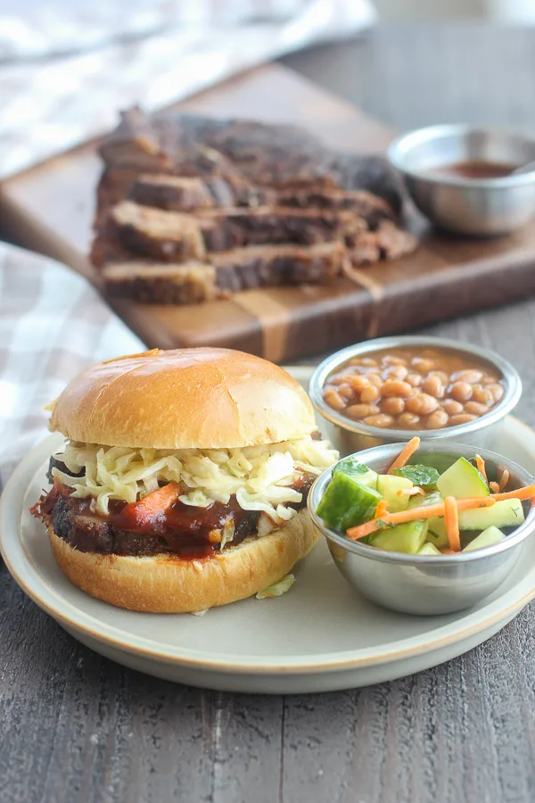 Slow cooker brisket sandwich served with cucumber salad and baked beans, with sliced brisket in the background on a wooden board