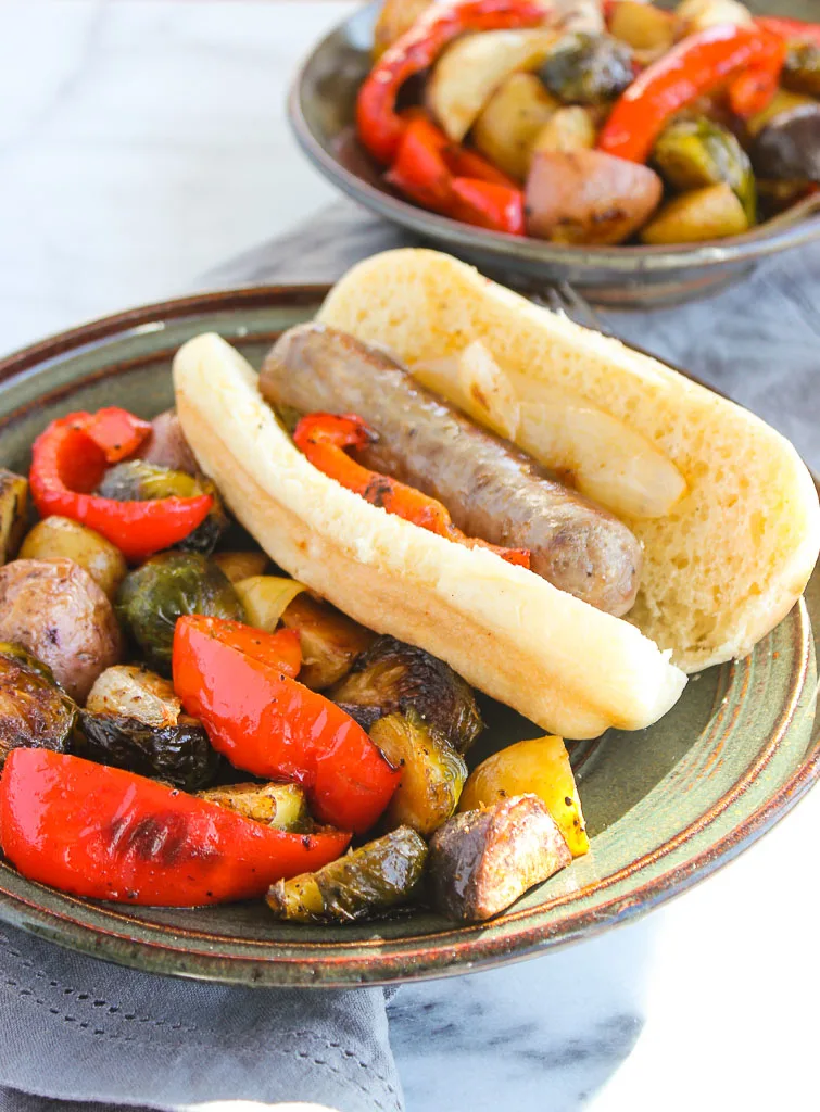 A roasted bratwurst served in a bun with roasted vegetables plated next to it.