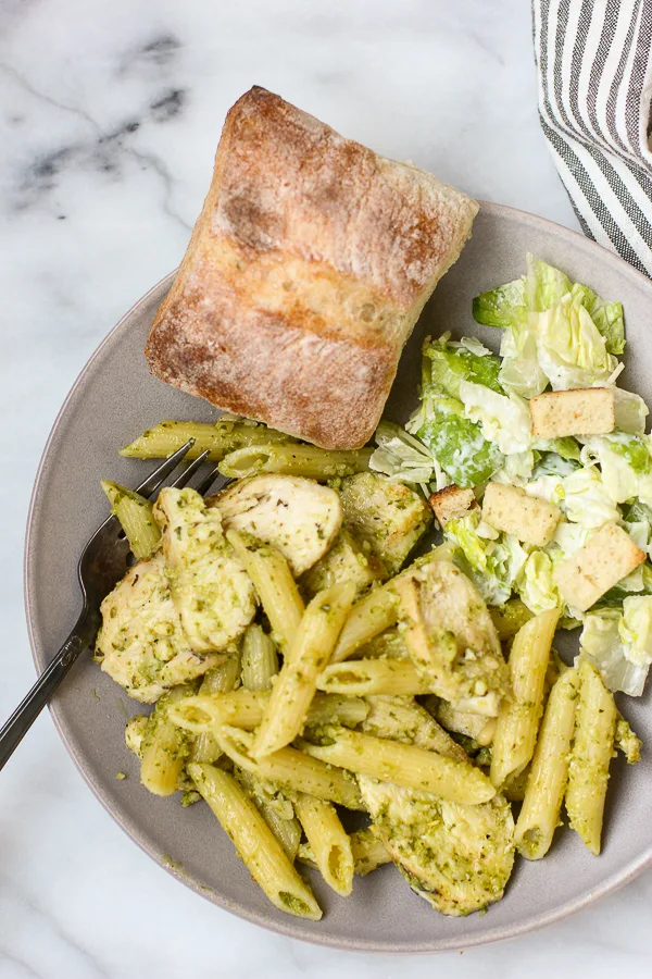Close up overhead photo of Chicken Pesto Pasta plated with caesar salad and a ciabatta roll