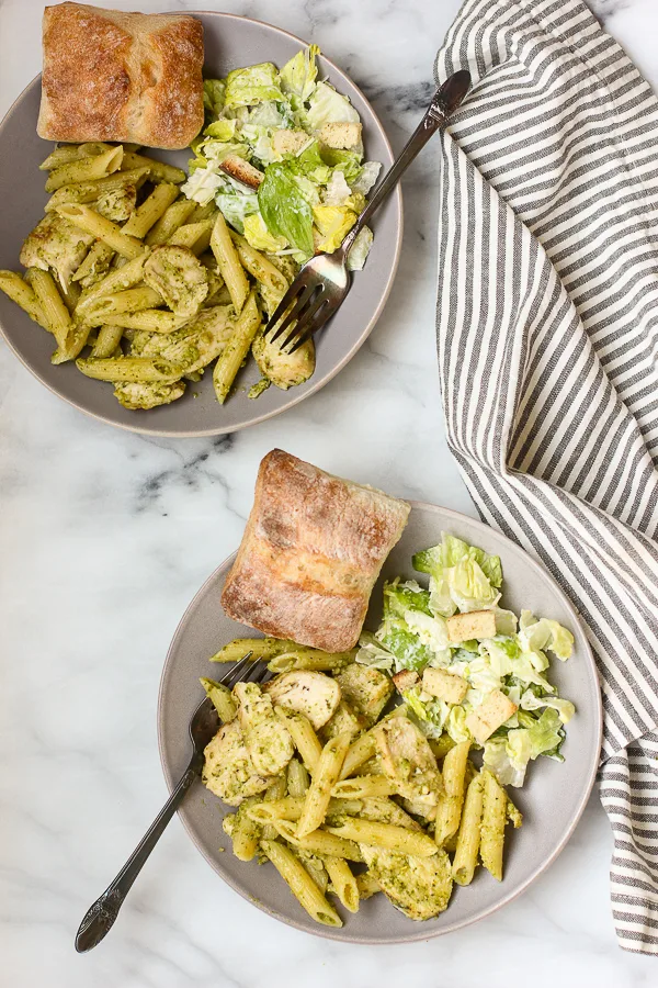 Overhead of two plates with grilled chicken pesto pasta, caesar salad and a ciabatta roll