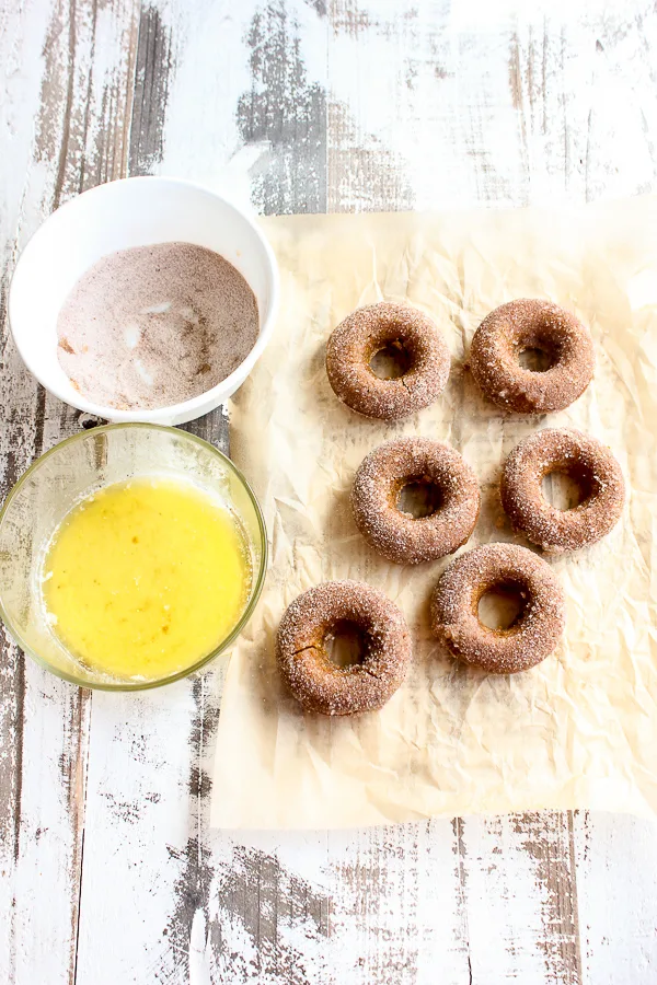 Baked Pumpkin donuts on parchment next to a bowl of cinnamon sugar and a bowl of melted butter