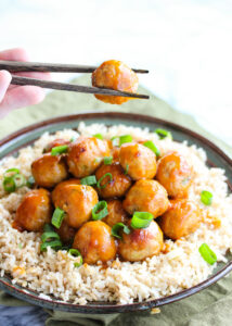 Orange chicken meatballs plated on a bed of rice with one meatball picked up with chopsticks
