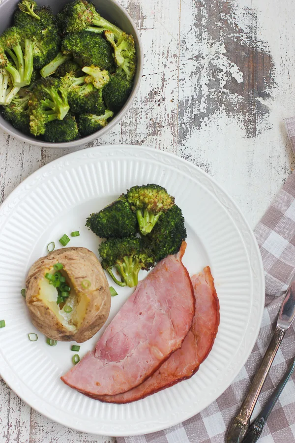 Roasted broccoli plated with ham slices and a baked potato, with the remaining broccoli in a serving bowl