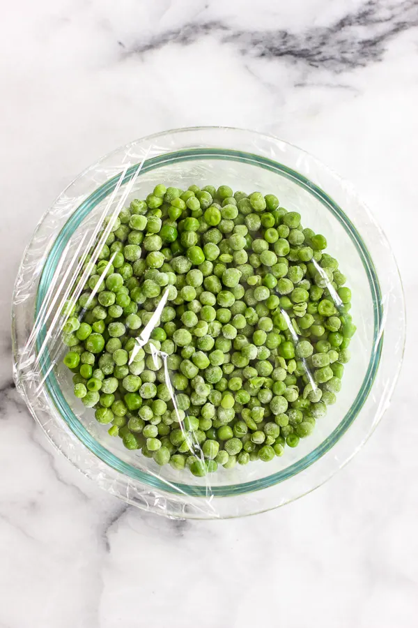 Frozen peas in a bowl covered with plastic wrap, ready to be microwaved