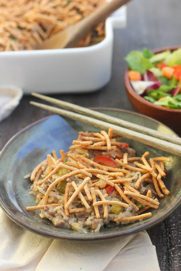 Chow mein casserole in a bowl with chopsticks served with mixed greens with the baking dish in the background